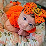 baby, infant, newborn, blue_eyes, orange_outfit, polka_dot_bow, halloween_bow, headband, pumpkin_decor, floral_pillow, cushion, closeup, portrait, lying_down, hands_together, cute, soft_lighting, warm_colors, fabric_pattern, props
