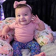 Joy joined the competition — help win amazing prizes! baby, infant, toddler, smiling, headband, pink_bow, pink_shirt, floral_pillow, patterned_leggings, barefoot, sitting, portrait, indoor, cozy_blanket, string_lights, curly_hair, blue_eyes, happy, earring, cute