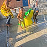 barefoot, blue_door, brick_wall, building, child, concrete, crocodile, diaper, happy, outdoor, playing, shorts, splash_pad, summer, sunlight, toddler, toy, water, water_splash, yellow_dress