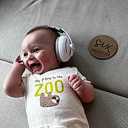 Adrian is registered to the contest to win money with this photo: baby, infant, headphones, smiling, couch, onesie, clothing, sloth, wooden_plaque, milestone, six_months, happy, portrait, indoor, child, cute, lying_down, face, expression, celebration