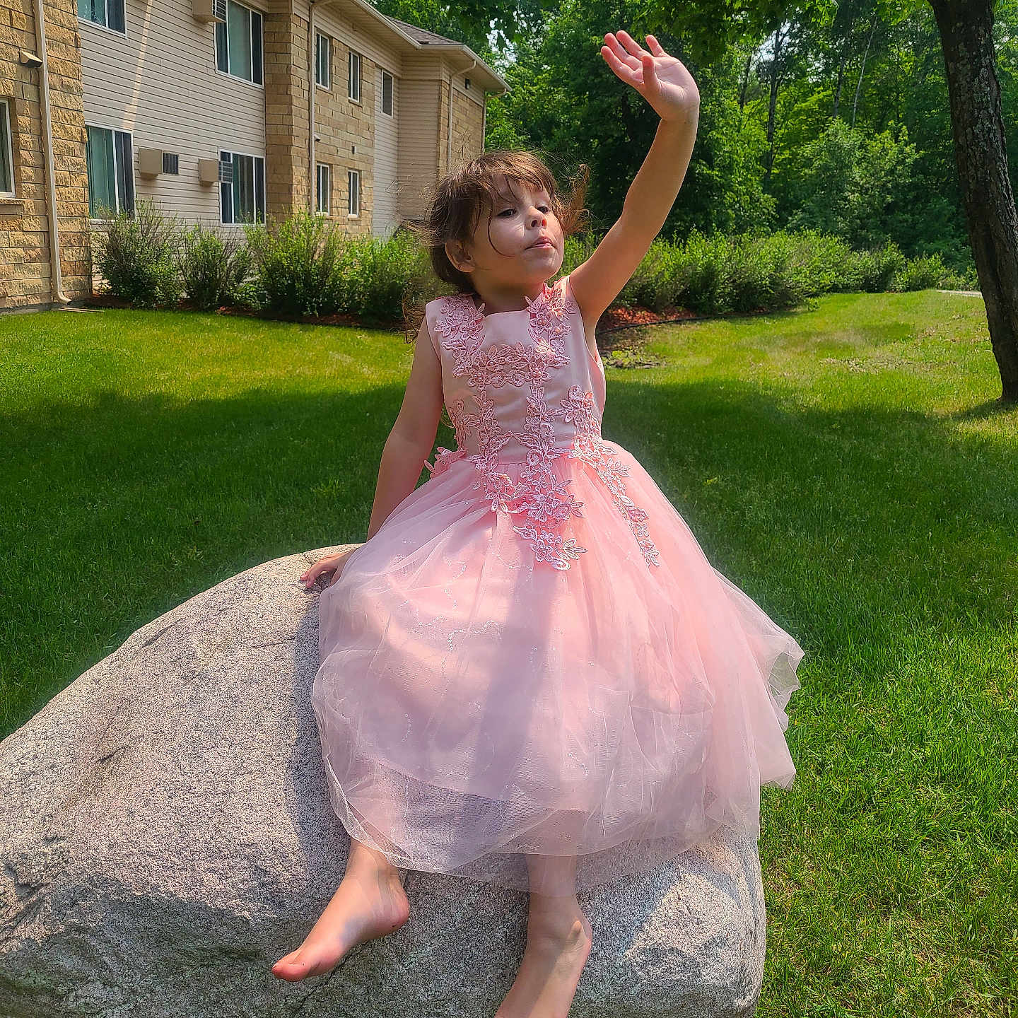 Davina is registered to the contest to win money with this photo: barefoot, building, bushes, casual, child, curly_hair, daylight, grass, greenery, nature, outdoor, pink_dress, playful, residential_area, rock, shadows, summer, sunlight, tree, waving