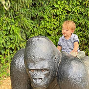 Noa a rejoint le concours — aidez-le/la à gagner de superbes lots ! toddler, child, statue, gorilla, outdoor, greenery, foliage, nature, curious, blond_hair, striped_shirt, casual_clothing, playful, sculpture, park, daylight, portrait, sitting, young_child, expression
