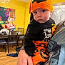 Iris is registered to the contest to win money with this photo: adult, baby, black_clothing, chair, child, expression, face, furniture, halloween_costume, hand, holding, home, indoor, living_room, orange_clothing, person, pumpkin_hat, table, wide_eyes, wood_floor