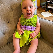Iris is registered to the contest to win money with this photo: baby, child, infant, smiling, green_dress, headband, bow, chair, cushion, indoors, happy, cute, person, portrait, hands, legs, toys, wooden_floor, keyboard, playroom