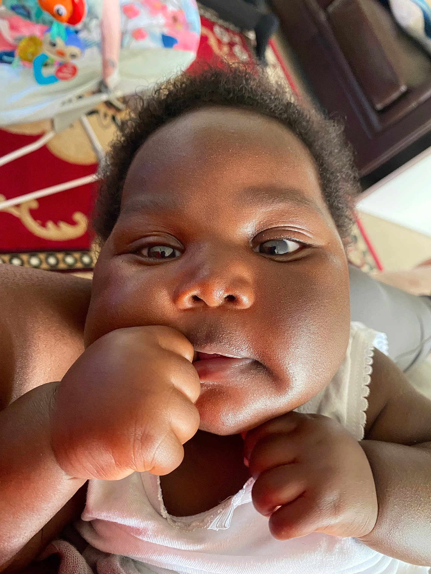 Gina is registered to the contest to win money with this photo: baby, close_up, face, hand, fist, curious, indoor, toy, colorful, skin, infant, child, white_clothing, expression, cheeks, person, young, cute, portrait, home