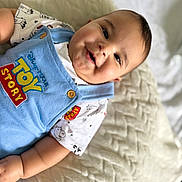 Enzo is registered to the contest to win money with this photo: baby, bed, blanket, blue_overalls, button, child, chubby_cheeks, closeup, clothing, cute, face, happy, indoors, infant, lying_down, patterned_shirt, smiling, soft_texture, toy_story, white_shirt