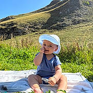 Azaan joined the competition — help win amazing prizes! baby, child, grass, hill, mountain, sunhat, blue_onesie, blanket, outdoor, nature, smiling, sitting, greenery, sunny, daylight, person, happy, field, rock, vegetation