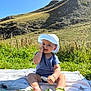 baby, child, grass, hill, mountain, sunhat, blue_onesie, blanket, outdoor, nature, smiling, sitting, greenery, sunny, daylight, person, happy, field, rock, vegetation