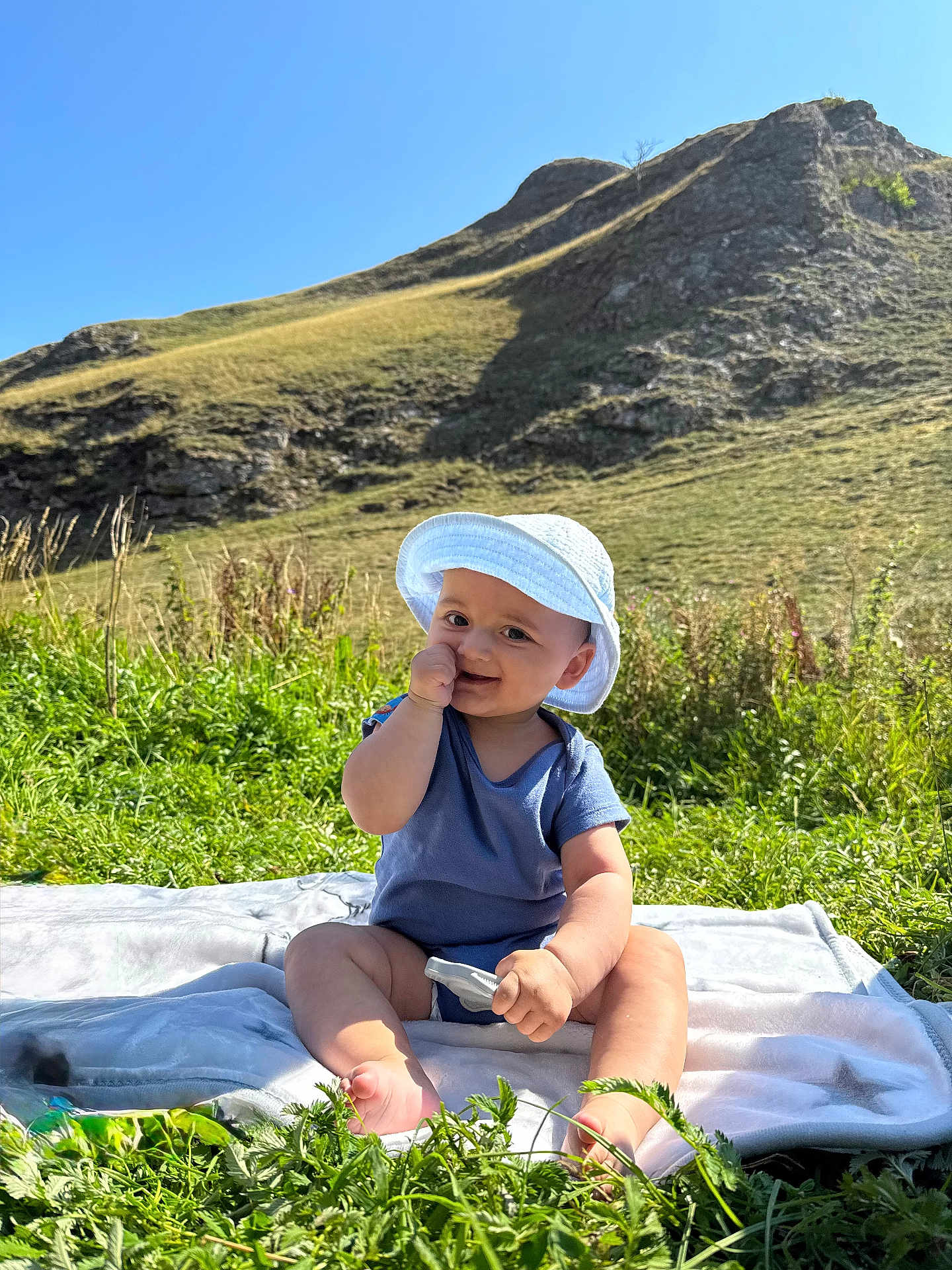 Azaan joined the competition — help win amazing prizes! baby, child, grass, hill, mountain, sunhat, blue_onesie, blanket, outdoor, nature, smiling, sitting, greenery, sunny, daylight, person, happy, field, rock, vegetation