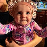 baby, child, infant, pink_bonnet, colorful_pajamas, cartoon_print, smiling_adult, kitchen, cabinet, countertop, fruit_basket, curious_expression, closeup, indoor, person, holding, cute, portrait, happy, family