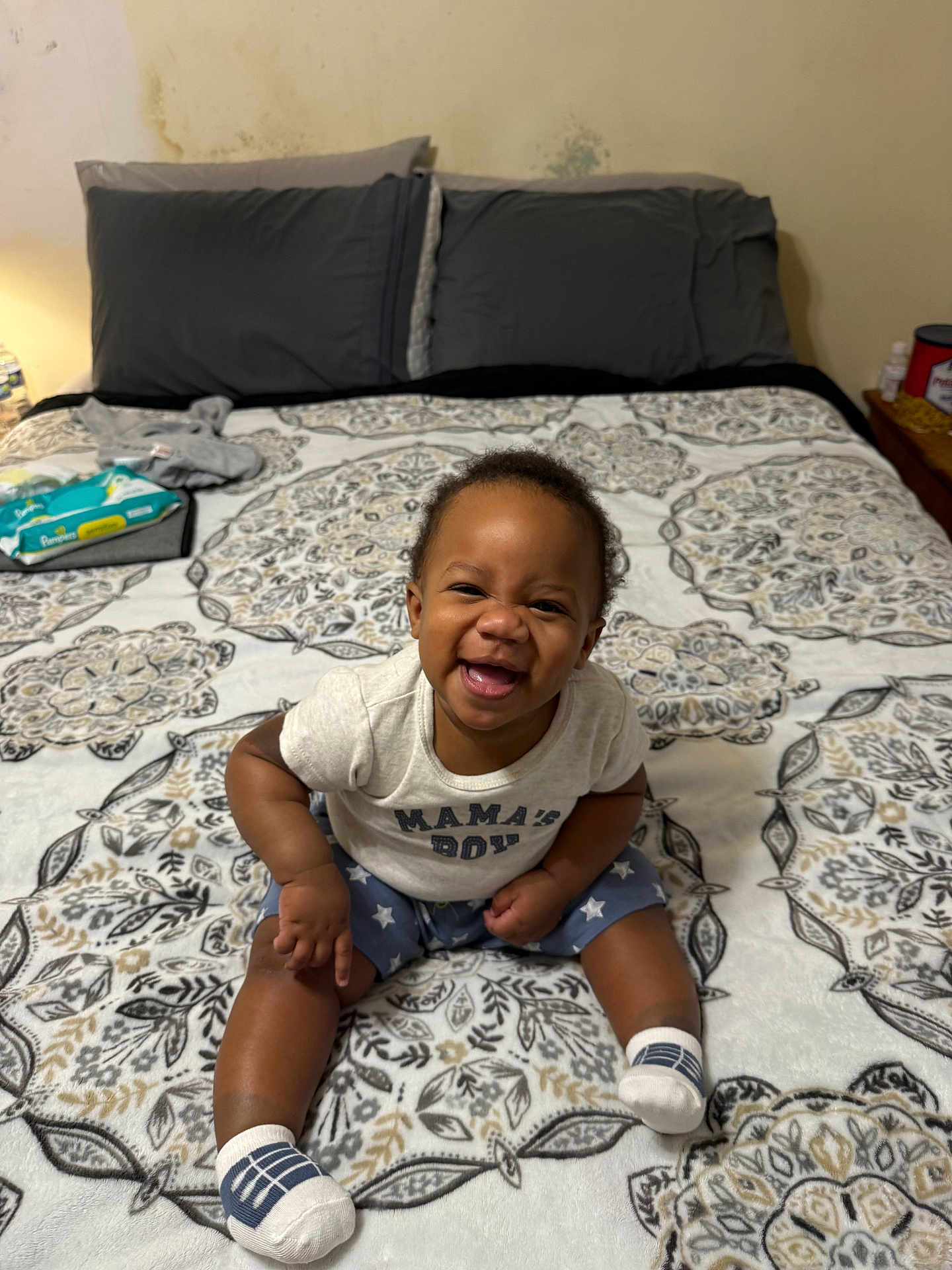 Christian is registered to the contest to win money with this photo: baby, child, smiling, sitting, bed, pillow, blanket, pattern, clothing, socks, indoor, room, joyful, happy, person, face, infant, toddler, text_on_clothing, furniture