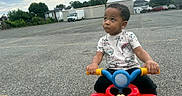 Jayse is registered to the contest to win money with this photo: asphalt, buildings, casual_clothing, child, cloudy_sky, curly_hair, daytime, dinosaur_print, fence, hands_on_handlebars, looking_up, mickey_mouse, outdoor, parking_lot, ride_on_toy, shirt, toddler, toy, trees, vehicle
