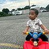 Jayse is registered to the contest to win money with this photo: toddler, child, toy, mickey_mouse, ride_on_toy, parking_lot, outdoor, dinosaur_print, shirt, curly_hair, casual_clothing, vehicle, asphalt, cloudy_sky, trees, buildings, fence, daytime, looking_up, hands_on_handlebars