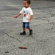 Jayse is registered to the contest to win money with this photo: child, boy, mickey_mouse, tshirt, denim_shorts, black_shoes, pinecone, asphalt, cracks, outdoor, parking_lot, car, grass, tree, playful, walking, smiling, person, casual_clothing, daytime