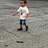 child, boy, mickey_mouse, tshirt, denim_shorts, black_shoes, pinecone, asphalt, cracks, outdoor, parking_lot, car, grass, tree, playful, walking, smiling, person, casual_clothing, daytime