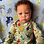 Saint is registered to the contest to win money with this photo: baby, child, blue_eyes, curly_hair, dinosaur_pajamas, pajamas, blanket, soft_texture, indoor, portrait, infant, cute, seated, clothing, person, closeup, expression, hands, face, nursery