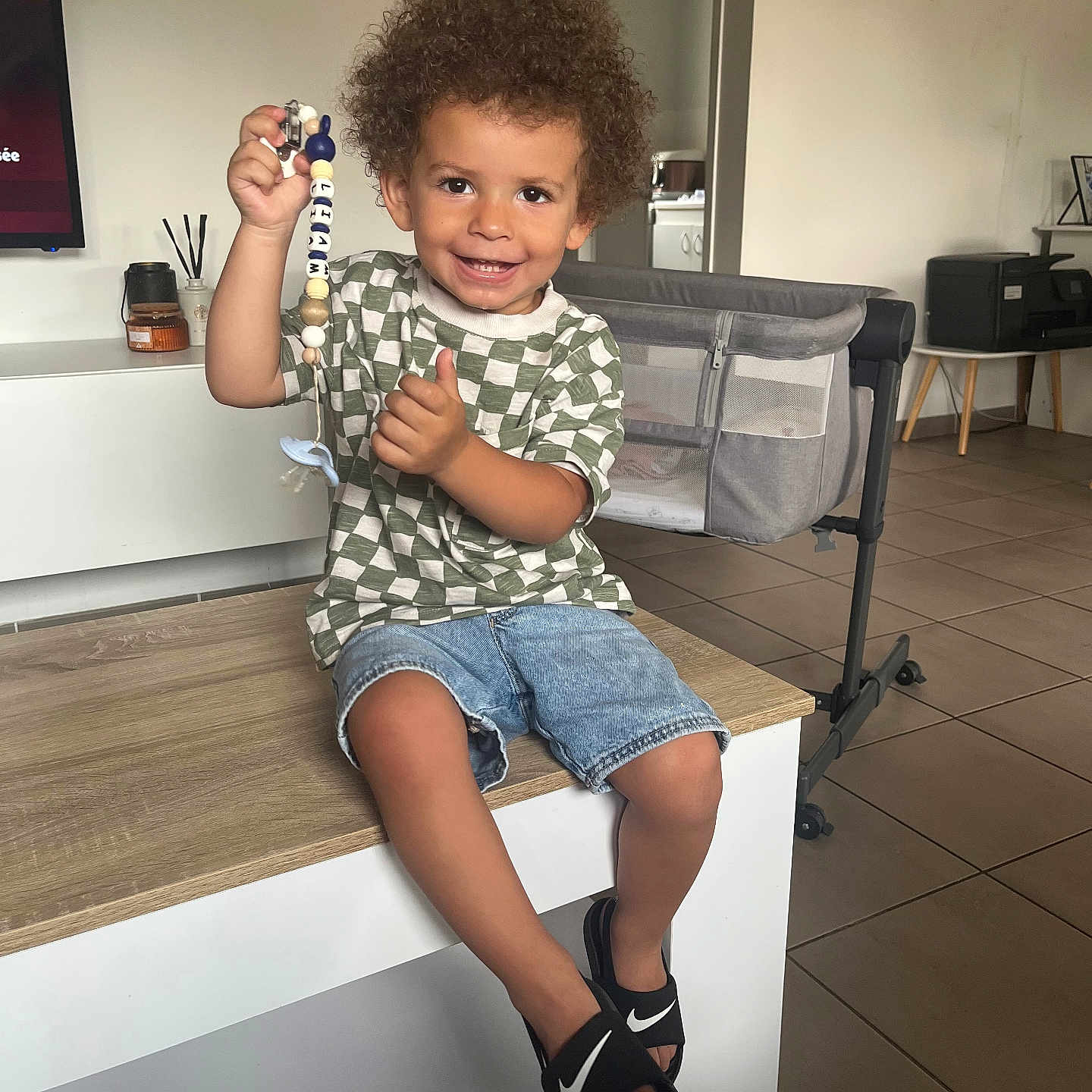 Liam a rejoint le concours — aidez-le/la à gagner de superbes lots ! bassinet, beads, checkered_shirt, child, curly_hair, cute, denim_shorts, footwear, furniture, happy, home, indoor, living_room, nike_slides, pacifier_clip, person, portrait, smiling, table, tiled_floor