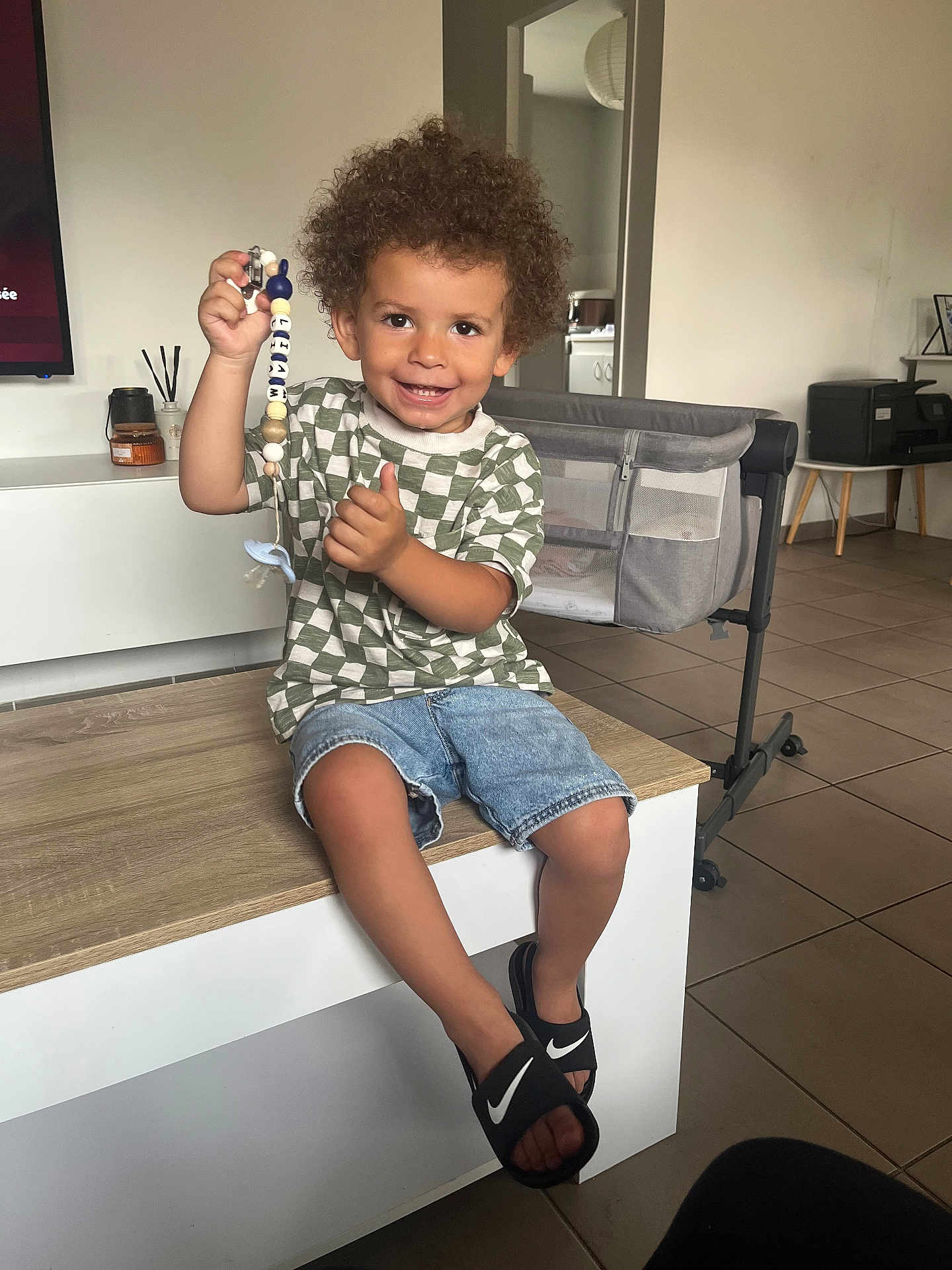 Liam a rejoint le concours — aidez-le/la à gagner de superbes lots ! child, smiling, curly_hair, indoor, table, pacifier_clip, beads, denim_shorts, tiled_floor, living_room, bassinet, footwear, nike_slides, checkered_shirt, person, happy, furniture, home, cute, portrait