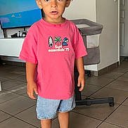Liam participe au concours pour gagner de l'argent avec cette photo : child, curly_hair, indoor, tiled_floor, pink_tshirt, denim_shorts, sneakers, white_socks, television, cartoon, bassinet, playpen, standing, curious_expression, casual_clothing, young_boy, home_interior, person, footwear, modern