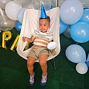 Ethan Marvin participe au concours pour gagner de l'argent avec cette photo : toddler, child, party_hat, balloons, chair, smiling, happy, celebration, indoor, footwear, shorts, striped_shirt, green_carpet, plastic_chair, party_horn, blue, white, birthday, festive, cute