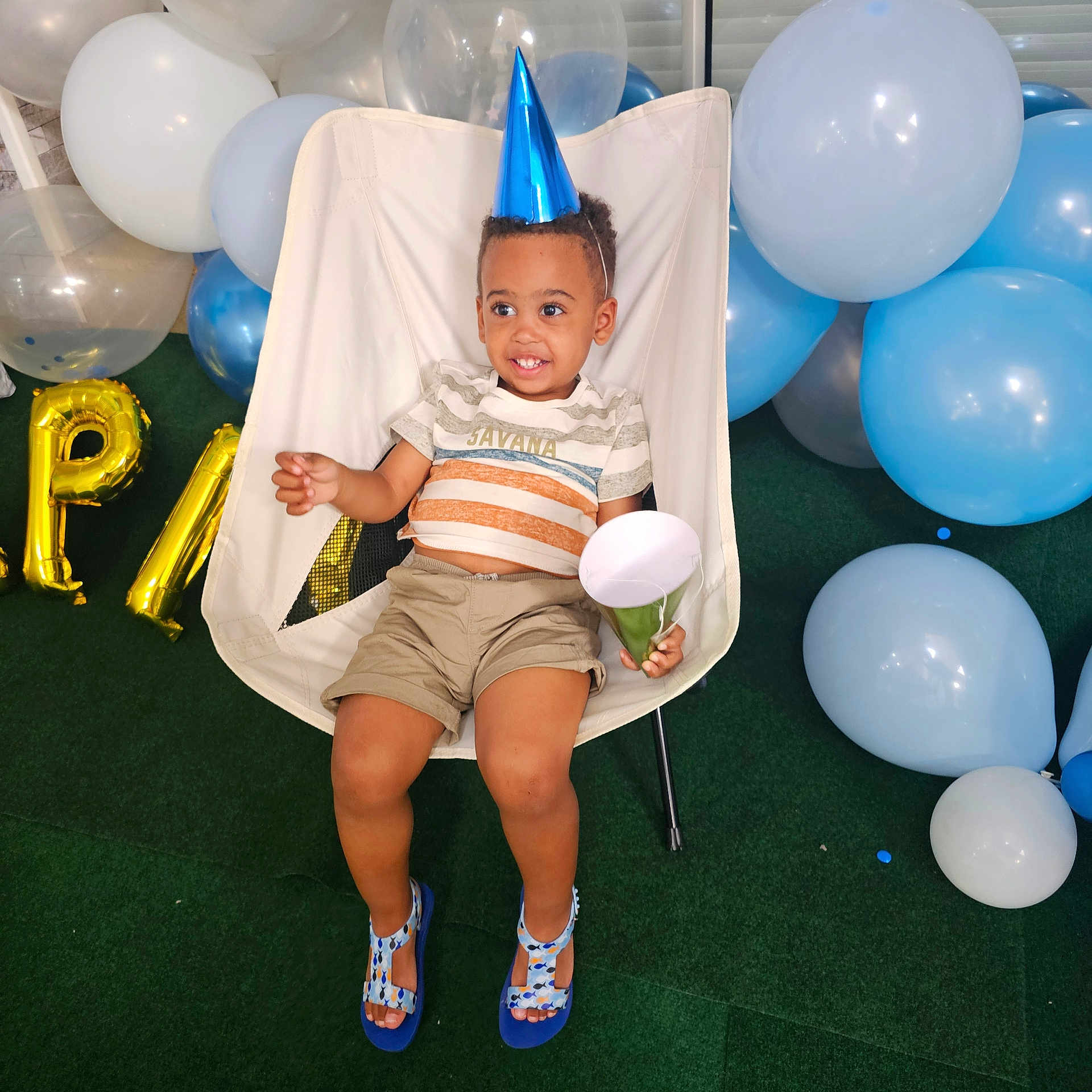 Ethan Marvin participe au concours pour gagner de l'argent avec cette photo : toddler, child, party_hat, balloons, chair, smiling, happy, celebration, indoor, footwear, shorts, striped_shirt, green_carpet, plastic_chair, party_horn, blue, white, birthday, festive, cute
