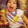 award, baby, big_eyes, celebration, champion, child, confetti, cute, excited, glitter, gold_medal, hair_ties, holding_trophy, party, portrait, sash, smiling, toddler, trophy, white_tshirt