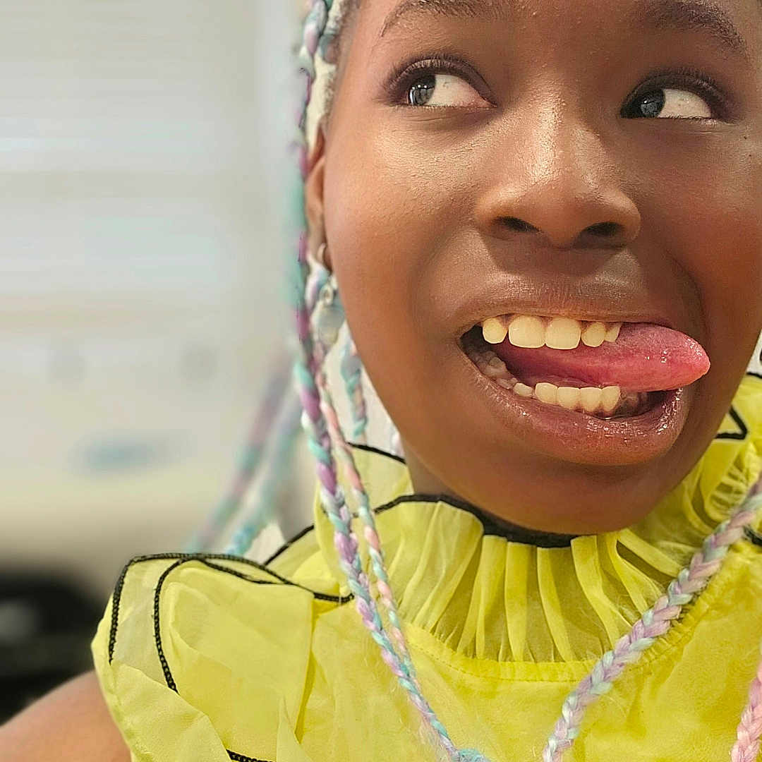 Amari participe au concours pour gagner de l'argent avec cette photo : braided_hair, casual, child, closeup, colorful_hair, expression, face, fashion, fun, happy, indoor, natural_light, person, playful, portrait, ruffled_top, smiling, tongue, yellow_clothing, young_person