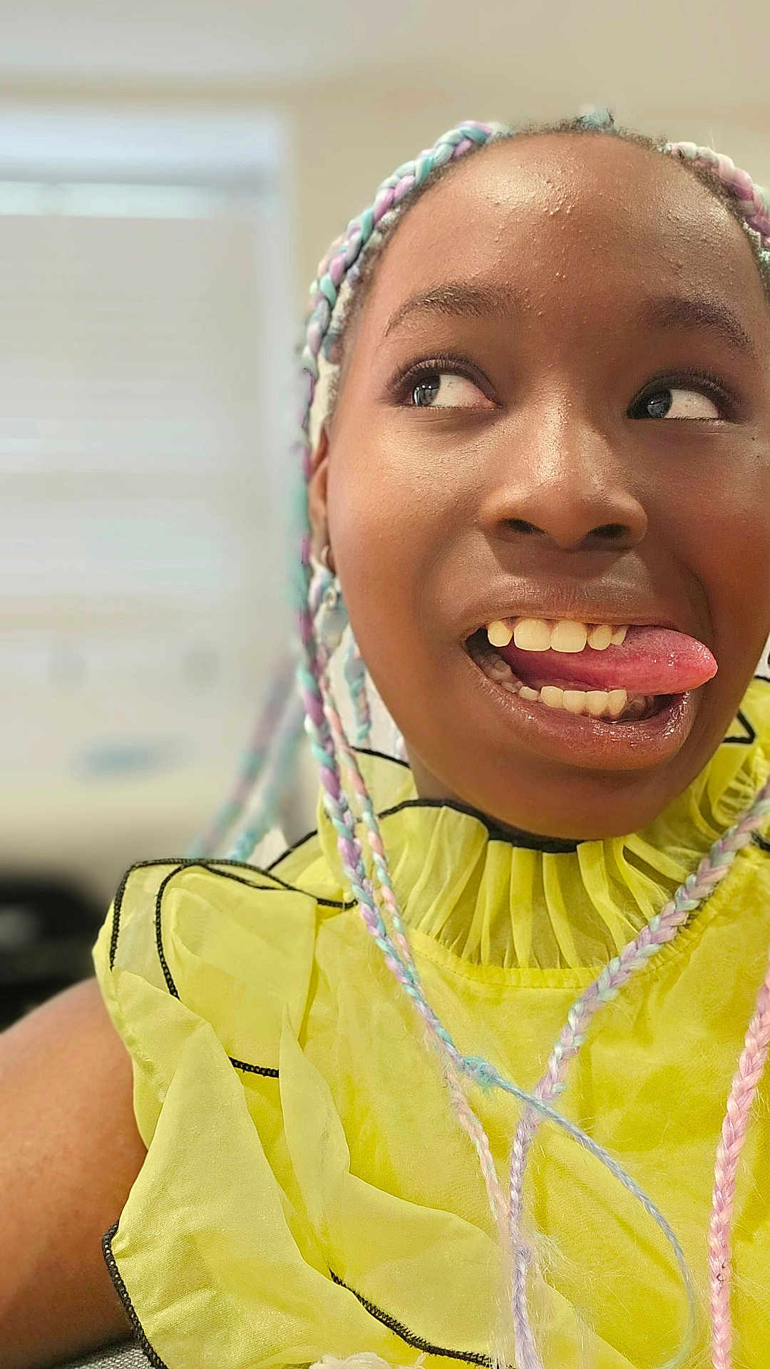 Amari participe au concours pour gagner de l'argent avec cette photo : child, face, tongue, braided_hair, colorful_hair, yellow_clothing, ruffled_top, indoor, portrait, playful, young_person, smiling, expression, natural_light, closeup, person, fashion, happy, fun, casual
