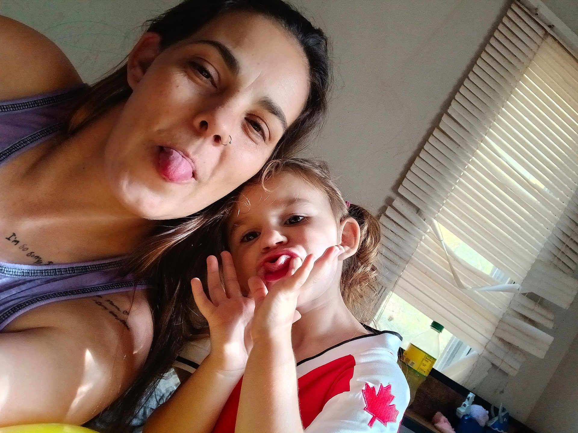 Maddie is registered to the contest to win money with this photo: woman, child, indoor, window, blinds, tongue_out, playful, selfie, face, pucker, hair, shirt, canada_flag, tattoo, light, expression, person, casual, fun, family