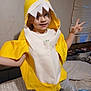 Maddie is registered to the contest to win money with this photo: child, yellow_costume, shark_costume, bed, blanket, indoor, window, blinds, smiling, playful, person, pajamas, bedroom, furniture, toy, curtains, happy, posing, casual, home