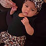 baby, infant, headband, leopard_print, black_clothing, hand, adult_hand, holding, lying_down, cute, calm, curious, face, person, skin, indoor, soft_lighting, patterned_pants, closeup, portrait