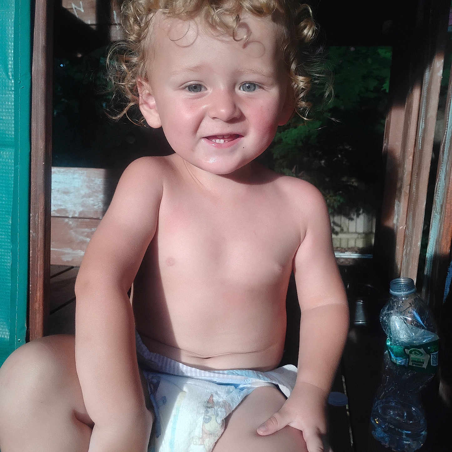 Douglas joined the competition — help win amazing prizes! toddler, child, curly_hair, diaper, smile, pointing, sunlight, wooden_structure, water_bottle, outdoor, skin, bare_chest, playful, young_child, person, face, sitting, cute, daylight, expression