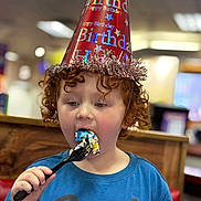 Shane is registered to the contest to win money with this photo: child, birthday, party_hat, cake, fork, curly_hair, blue_shirt, indoor, celebration, eating, food, happy, red_hat, closeup, person, fun, colorful, smile, face, curly
