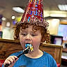 child, birthday, party_hat, cake, fork, curly_hair, blue_shirt, indoor, celebration, eating, food, happy, red_hat, closeup, person, fun, colorful, smile, face, curly