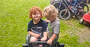 Shane is registered to the contest to win money with this photo: child, toddler, toy_car, curly_hair, grass, bicycle, outdoor, play, greenery, smile, fun, summer, park, vehicle, garden, happy, person, casual_clothing, daylight, nature