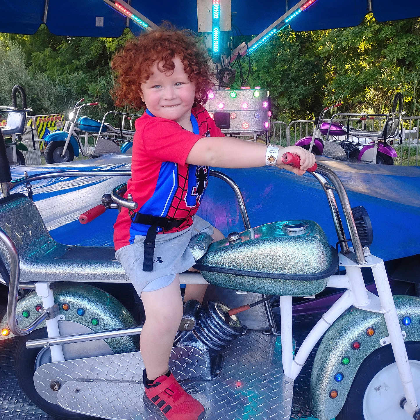 Shane joined the competition — help win amazing prizes! child, red_hair, curly_hair, smile, motorcycle_ride, carnival, outdoor, blue_canopy, neon_lights, greenery, shorts, red_shoes, ride, metal_platform, amusement_ride, fun, playful, daylight, person, park