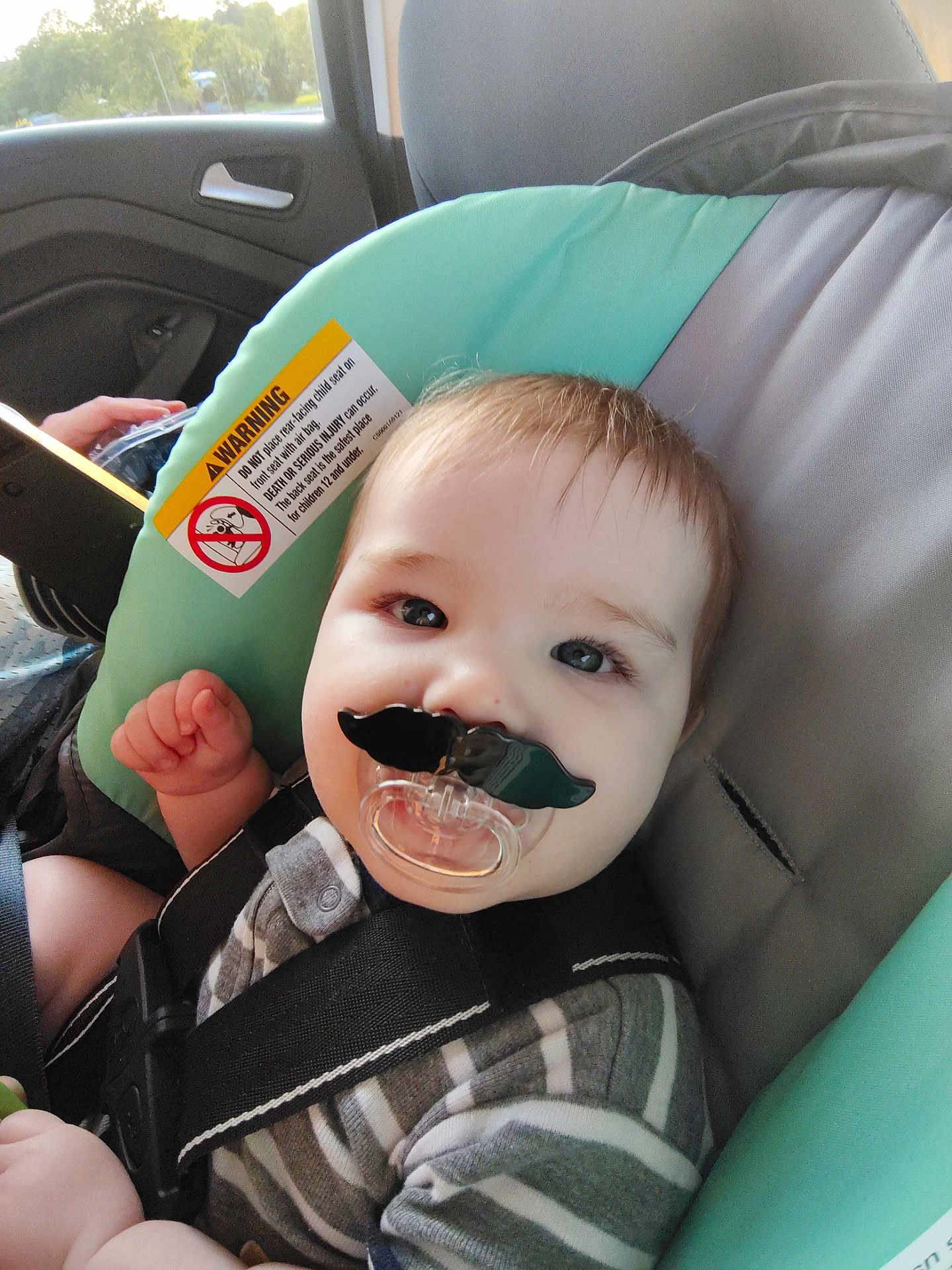 Liam joined the competition — help win amazing prizes! baby, car_seat, pacifier, mustache, child, striped_clothing, seatbelt, indoor, car, infant, cute, face, smiling, young_child, person, seat, warning_label, hand, hair, eyes