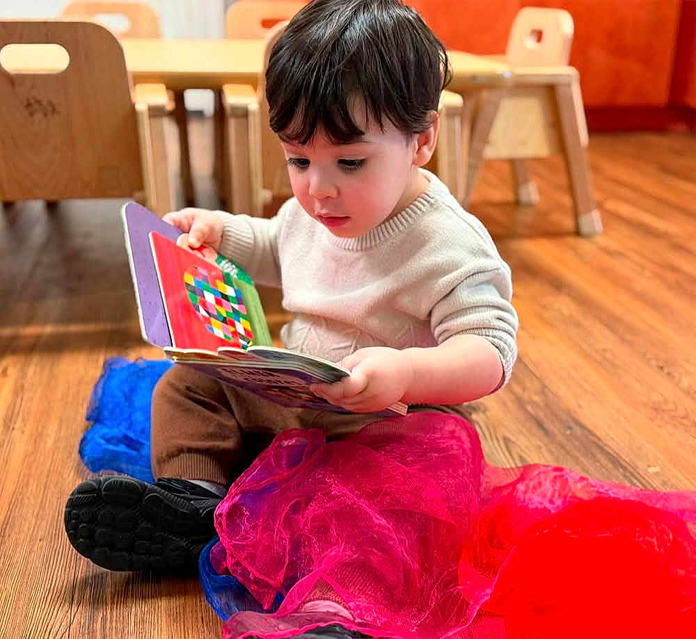 Ethan joined the competition — help win amazing prizes! toddler, child, reading, book, floor, wooden_floor, indoor, furniture, chair, table, colorful, fabric, red, blue, clothing, sitting, curly_hair, casual, concentration, young_child