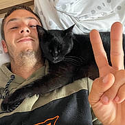 Switch participe au concours pour gagner de l'argent avec cette photo : man, cat, black_cat, pet, cuddling, sleeping, peace_sign, selfie, portrait, bed, bedroom, blanket, pillow, polka_dots, clothing, hoodie, logo, indoor, relaxed, human_face