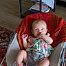 baby, infant, bouncer, thumb_sucking, colorful_clothing, indoor, carpet, table, books, floor, furniture, person, cute, child, home, relaxing, patterned_rug, wood_floor, small_table, baby_trend