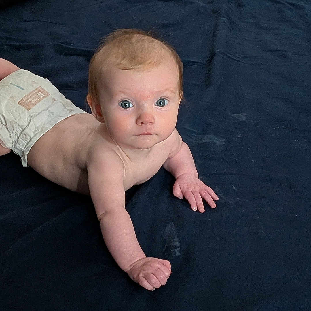 áine is registered to the contest to win money with this photo: baby, infant, diaper, blue_eyes, lying_down, blanket, pillow, indoor, curious, child, skin, young, sleeping_area, cozy, toddler, face, person, resting, cute, expression