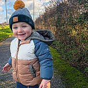 Eliott participe au concours pour gagner de l'argent avec cette photo : toddler, child, smiling, puffer_jacket, beanie, outdoor, pathway, hedge, moss, grass, cloudy_sky, sunlight, winter_clothing, happy, person, nature, casual, walking, blue_pants, shoes