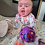 baby, child, infant, toy, stuffed_animal, elephant, ball, pacifier, blanket, polka_dot, floral_clothing, onesie, expression, indoor, carpet, face, hands, playing, cute, happy