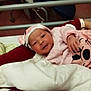 newborn, baby, infant, hospital_bed, person, pink_clothing, hat, blanket, arm, hospital_equipment, chair, indoor, bed, face, cute, lying_down, holding, smile, human, resting