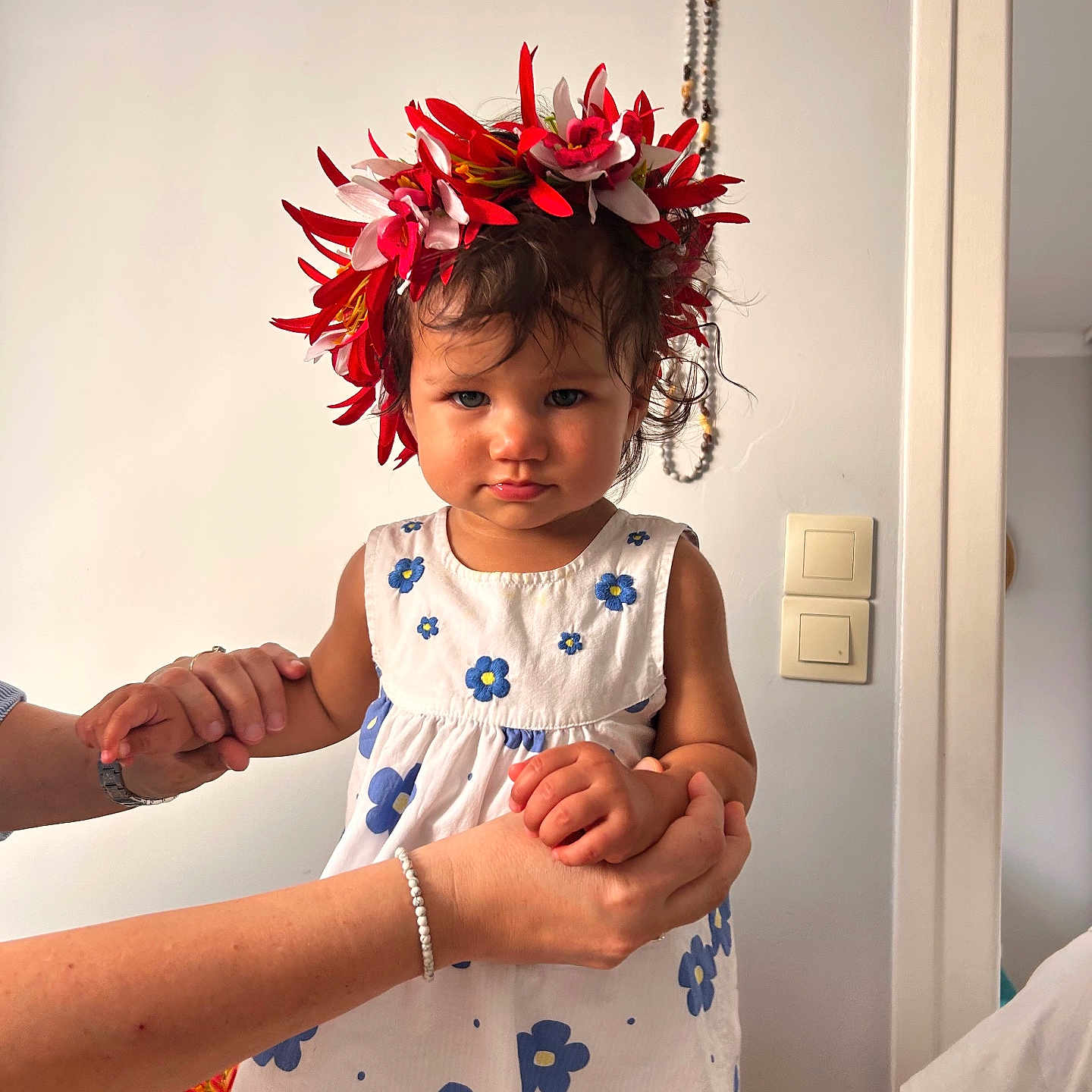 Ohaina participe au concours pour gagner de l'argent avec cette photo : adult_hands, beads, blue_flowers, bracelet, child, curly_hair, flower_crown, holding_hands, home, indoor, light_switch, person, portrait, serious_expression, skin, standing, toddler, wall, watch, white_dress