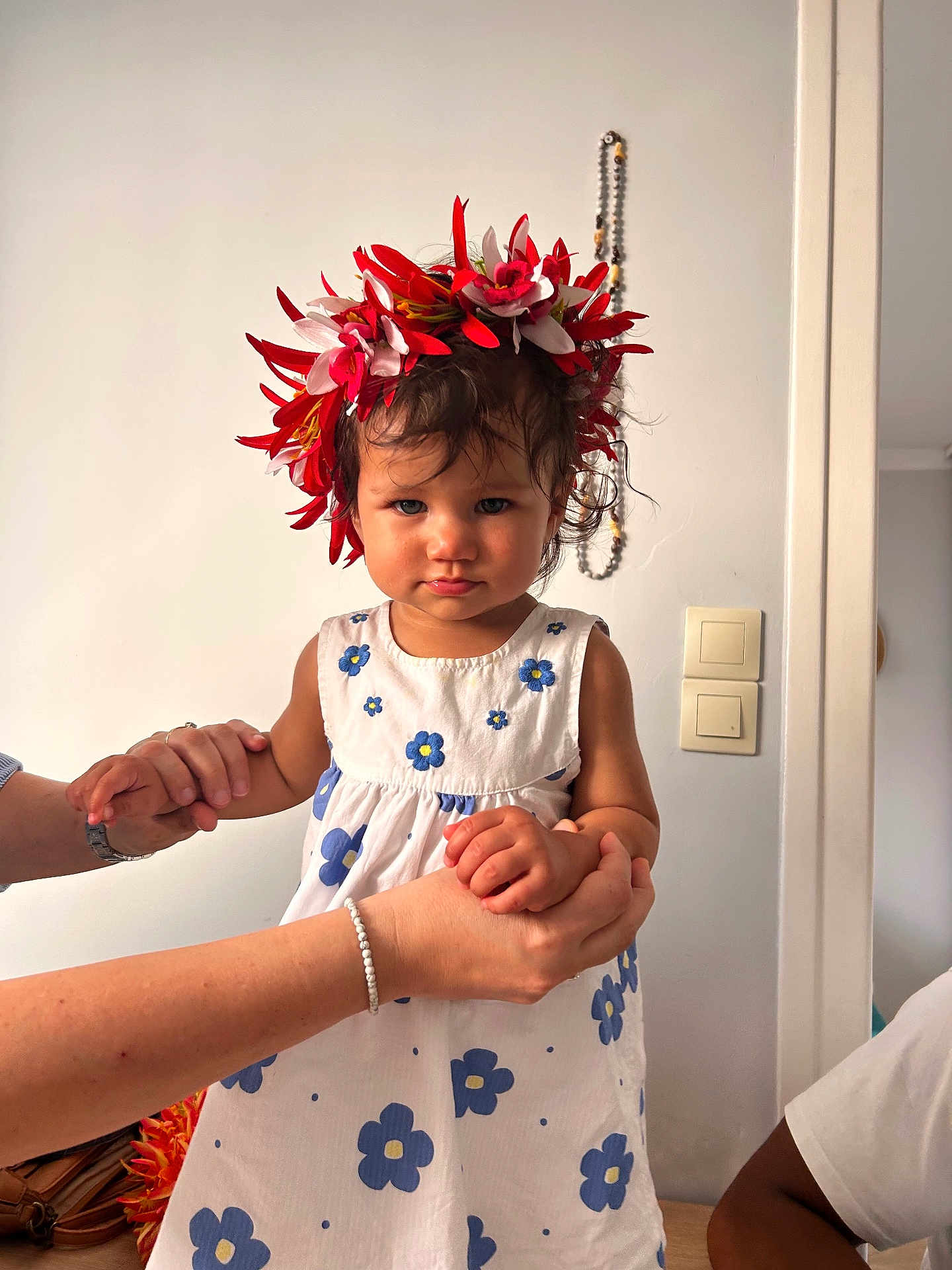 Ohaina participe au concours pour gagner de l'argent avec cette photo : child, toddler, flower_crown, white_dress, blue_flowers, indoor, holding_hands, adult_hands, curly_hair, serious_expression, beads, wall, light_switch, person, portrait, standing, skin, watch, bracelet, home