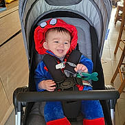 Ezio Jegu participe au concours pour gagner de l'argent avec cette photo : baby, child, stroller, costume, spiderman, red, blue, pacifier, indoor, floor, wooden_furniture, smiling, tongue_out, happy, seatbelt, toy, hood, cute, sitting, infant