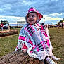 child, toddler, smiling, poncho, hood, outdoor, log, grass, cloudy_sky, people, event, footwear, shoes, happy, nature, evening, portrait, fringe, colorful_clothing, field