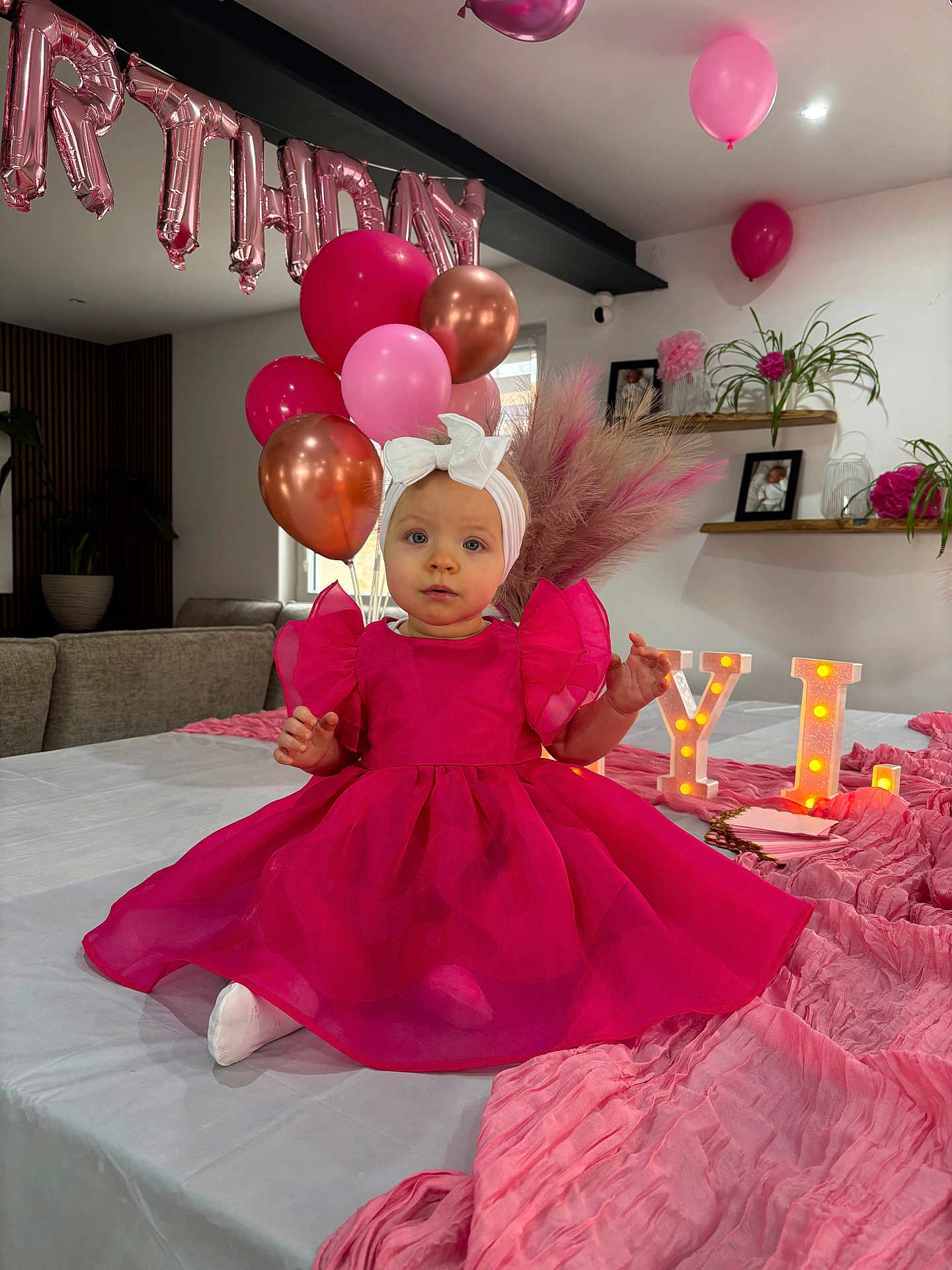 Lyla a rejoint le concours — aidez-le/la à gagner de superbes lots ! toddler, pink_dress, headband, balloons, birthday, table, marquee_letters, decorations, pink_theme, living_room, couch, plants, photos, celebration, party, fabric, indoor, child, smiling, festive