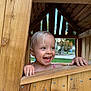 child, toddler, smiling, playhouse, wood, outdoor, hands, face, blue_eyes, short_hair, necklace, playground, natural_light, wooden_structure, happy, window, daylight, cute, person, joy